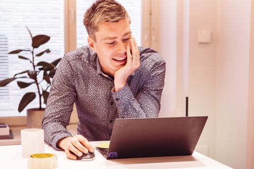 Mies istuu pöydän ääressä ja katsoo kannettavan tietokoneensa ruutua hymyillen.