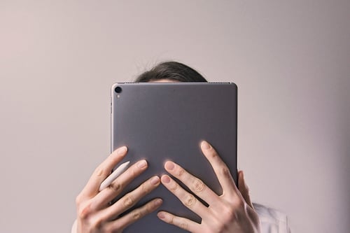 Nainen valkoisessa paidassa peittää kasvonsa tummanharmaalla iPad-tabletilla. 